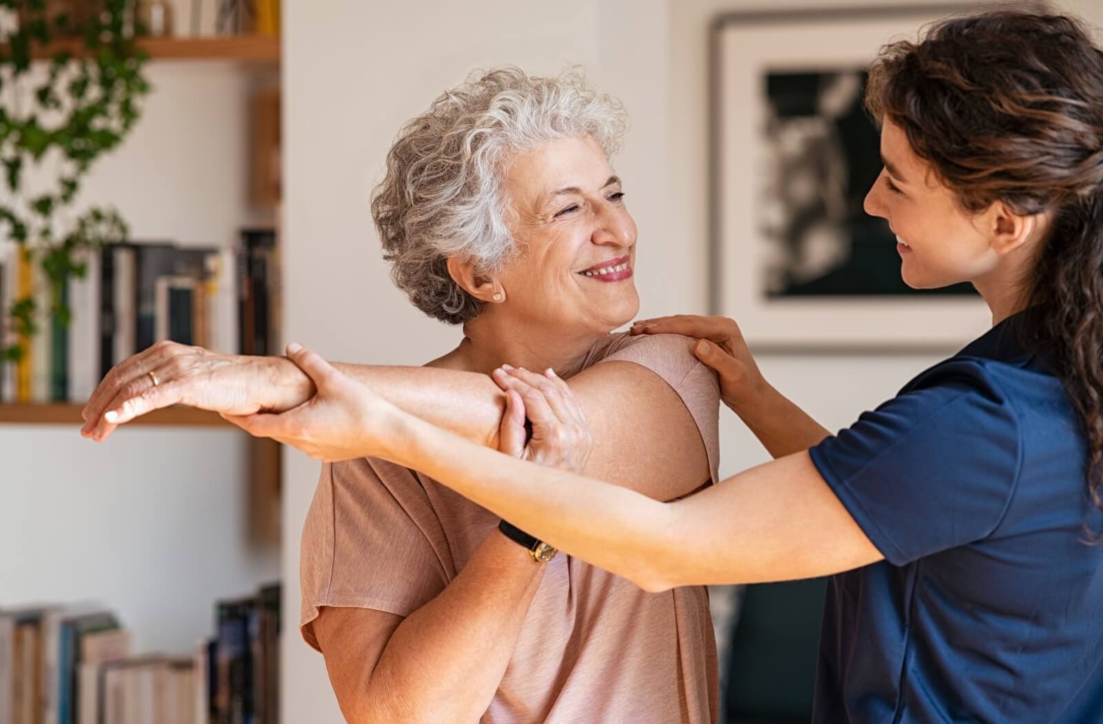 An elderly woman performing arm stretches at home under the guidance of a caregiver, as part of her post-acute brain injury rehabilitation session.