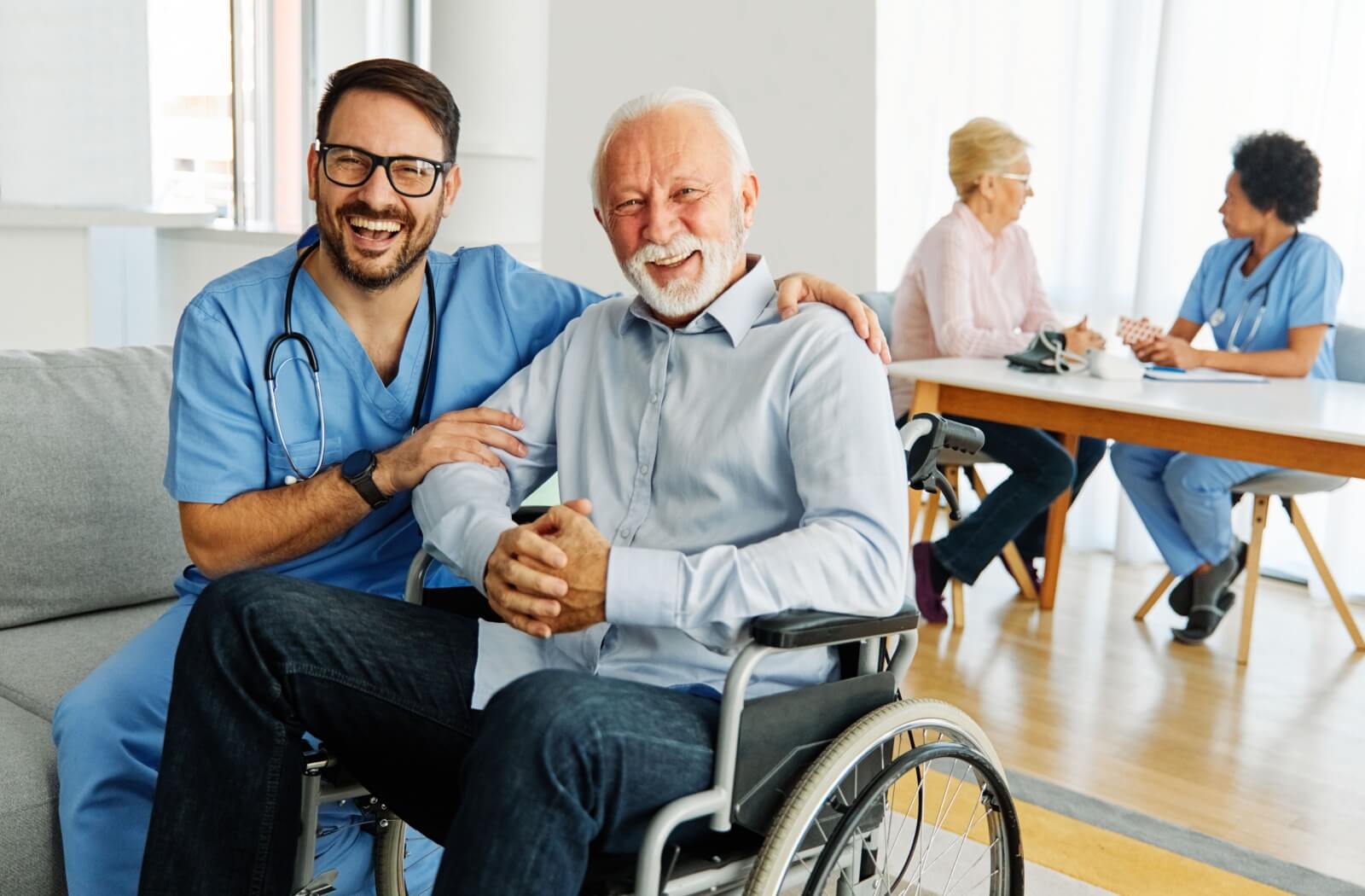 A caregiver sits on a couch next to an older adult in a wheelchair. They laugh together.