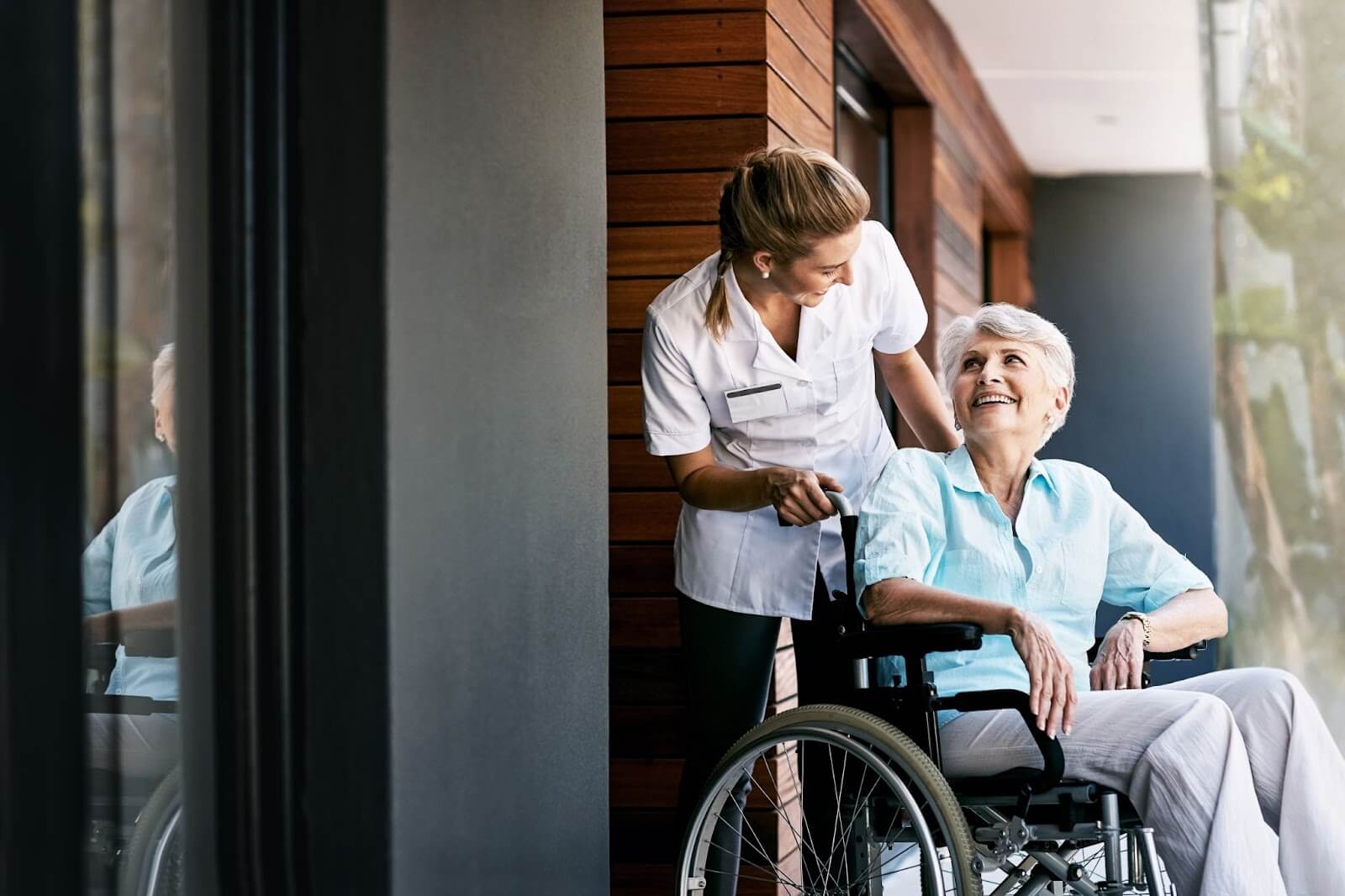A smiling nurse talks to an older adult woman in a wheelchair in a skilled nursing community.
