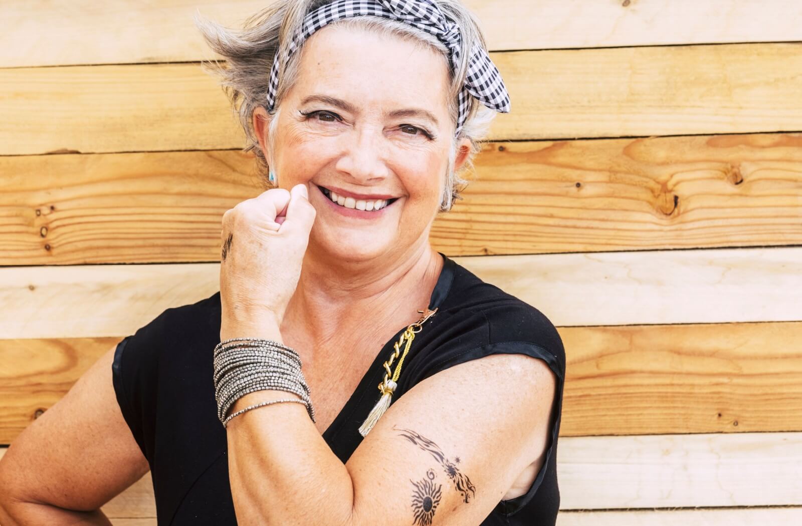 An older adult with short gray hair wears a headband and smiles while showing off their tattooed arm.