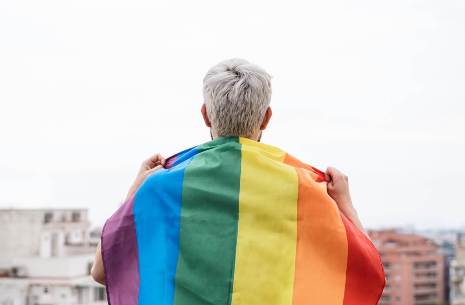 Older adult with short gray hair draped in a rainbow flag, standing on a rooftop overlooking the city.