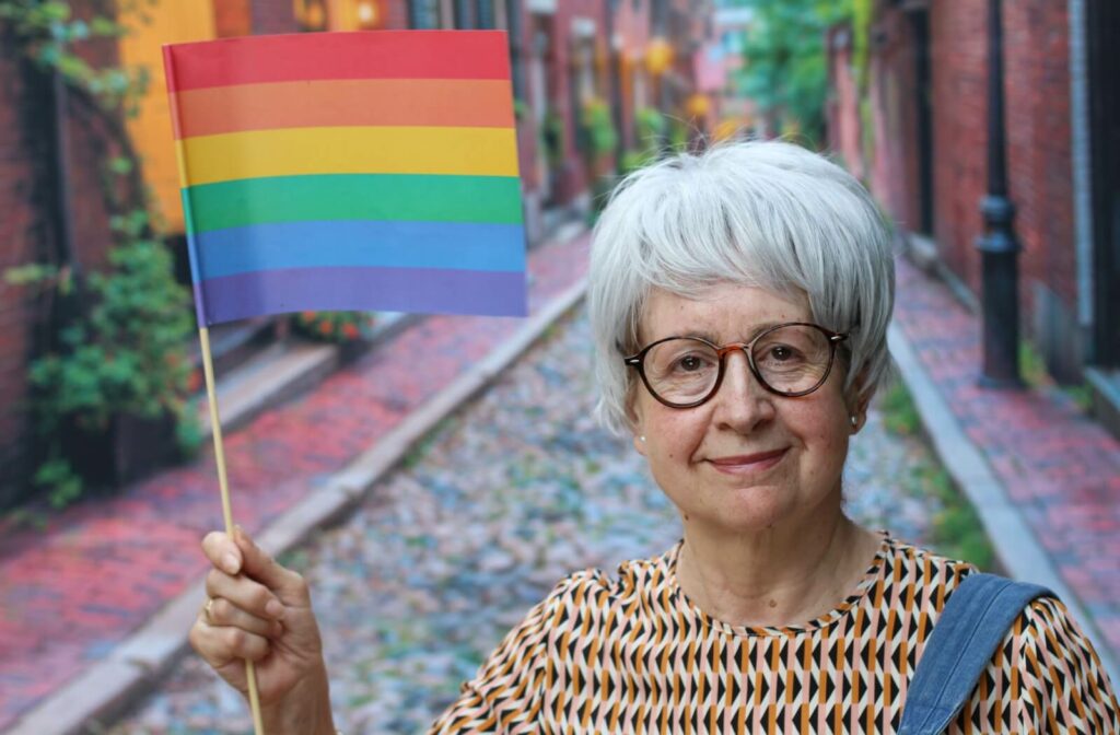 Older adult holding a rainbow flag on a cobblestone street, smiling at the camera.