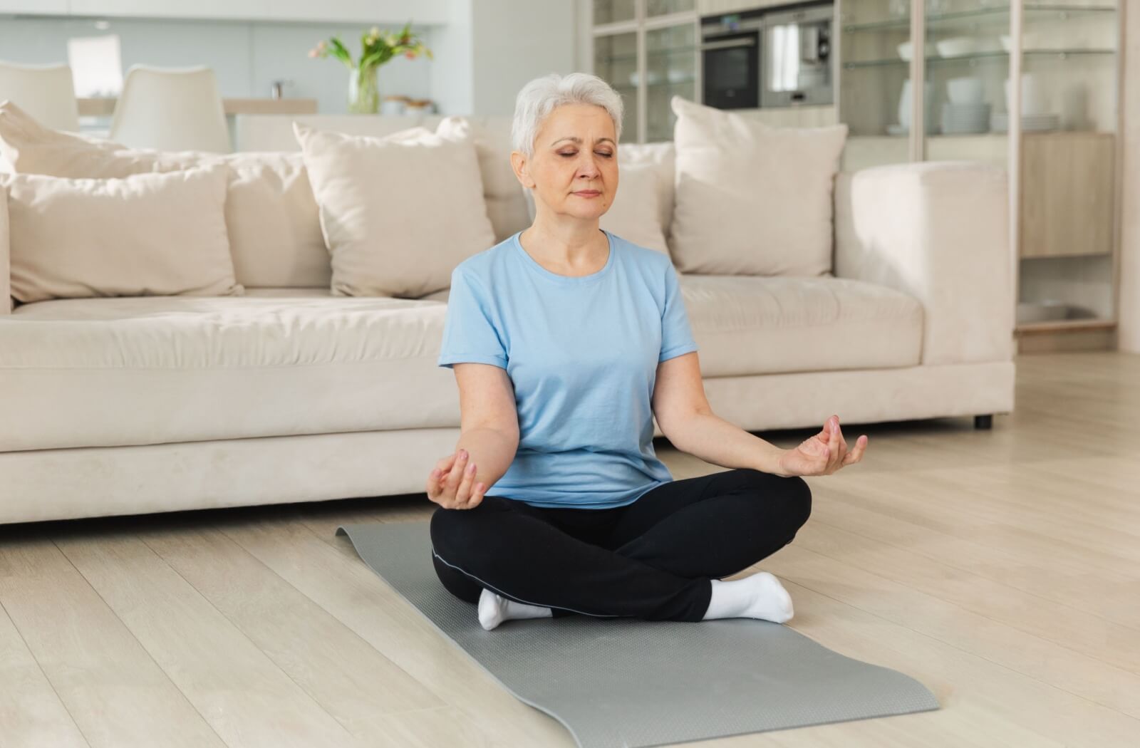 An older adult sits cross-legged on a yoga mat in their living room and meditates