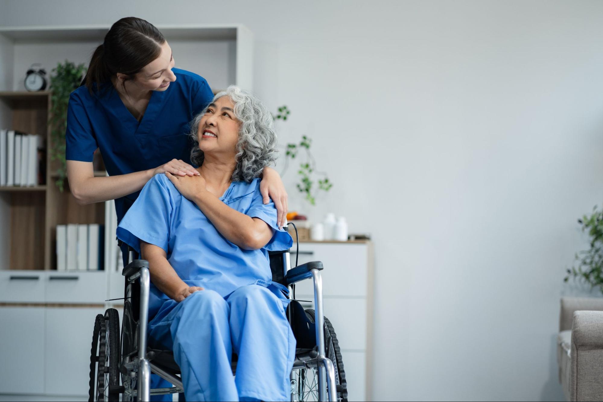 A smiling older adult woman in a wheelchair is comforted by an assisted living staff member.