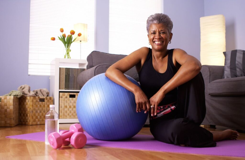 A senior sits happily on their yoga mat with an exercise ball and free weights after working out.