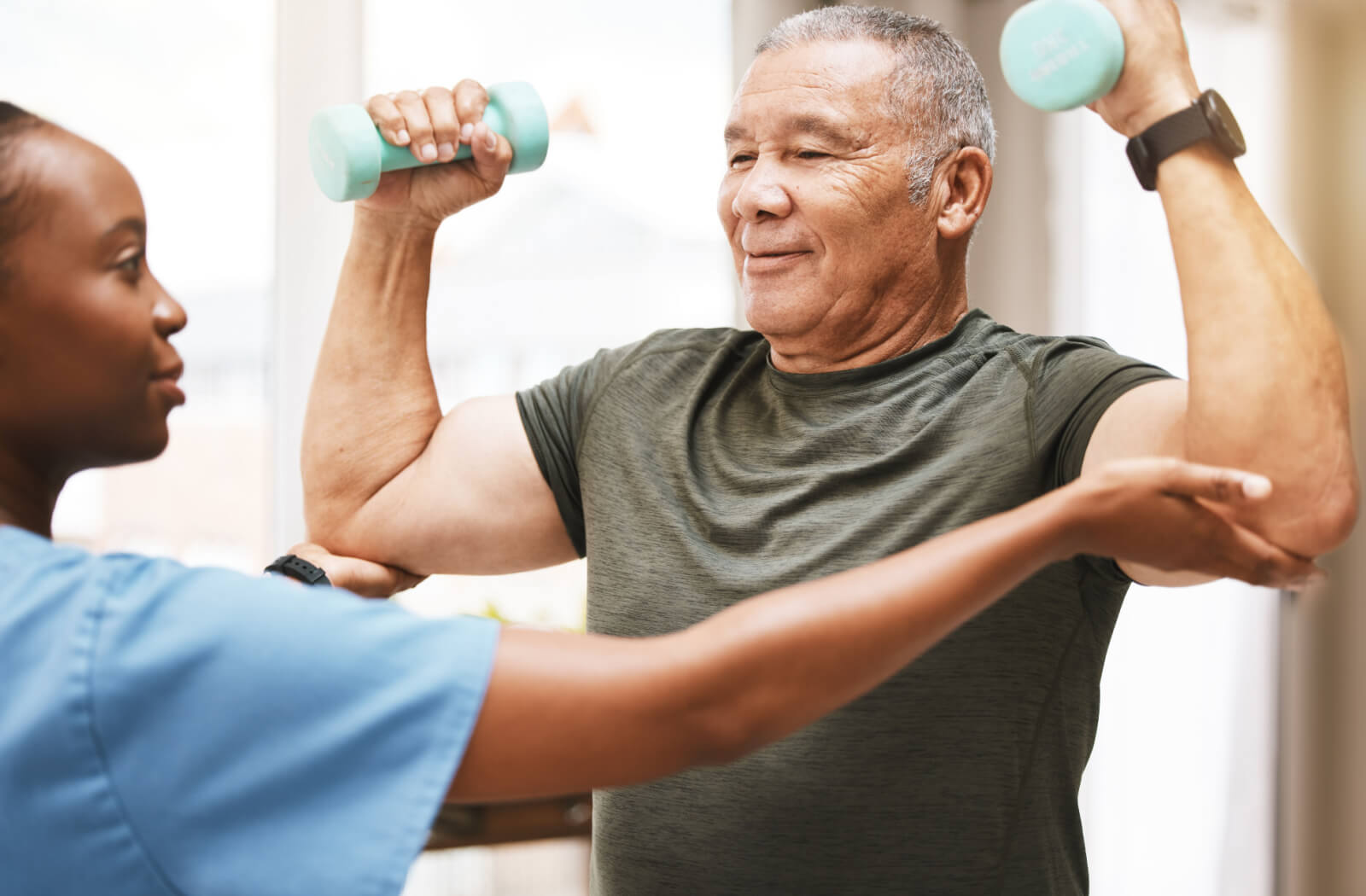 An older adult being assisted by a physical therapist to lift a pair of lightweight dumbbells.